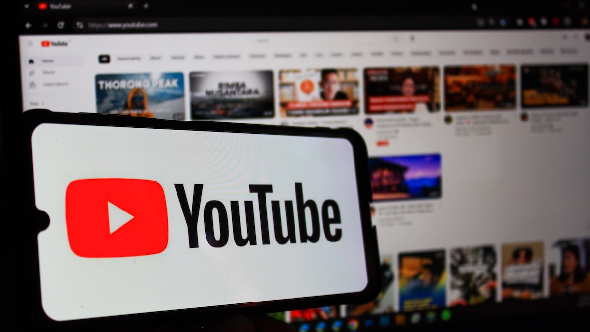 YouTube-Logo auf einem Smartphone, das vor einen Bildschirm gehalten wird, der eine Übersicht der Videoplattform zeigt