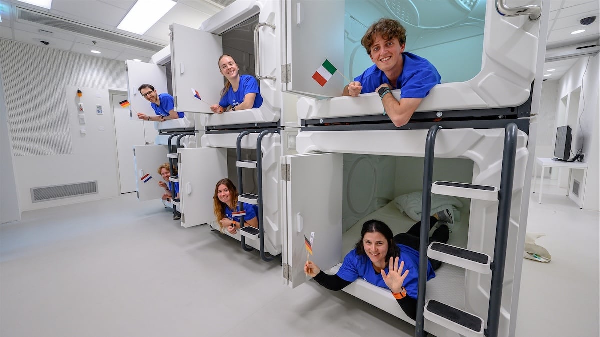 Studienteilnehmer für ein Isolationsexperiment: Sechs Menschen lächeln aus kleinen Schlafkapseln heraus und winken mit unterschiedlichen Landesfahnen