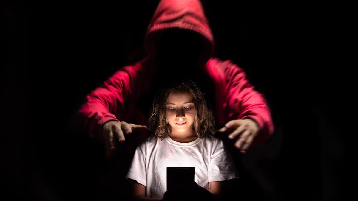 LKA warnt vor Taschengeld-Treffen: Mädchen steht im Dunkeln vor einem leuchtenden Smartphone, hinter ihr eine Gestalt mit Kapuze