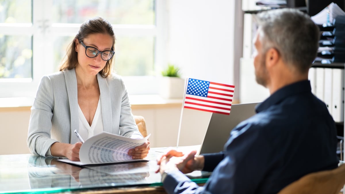 USA-Einreise nur noch mit Social-Media-Check: Regelungen sollen weiter verschärft werden 7 Interviewsituation bei der USA-Einreise: Mann mit USA-Fähnchen sitzt einer Frau gegenüber