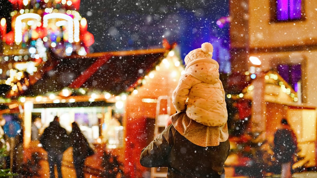 Kind sitzt auf den Schultern seines Vaters, der über einen hell erleuchteten Weihnachtsmarkt läuft