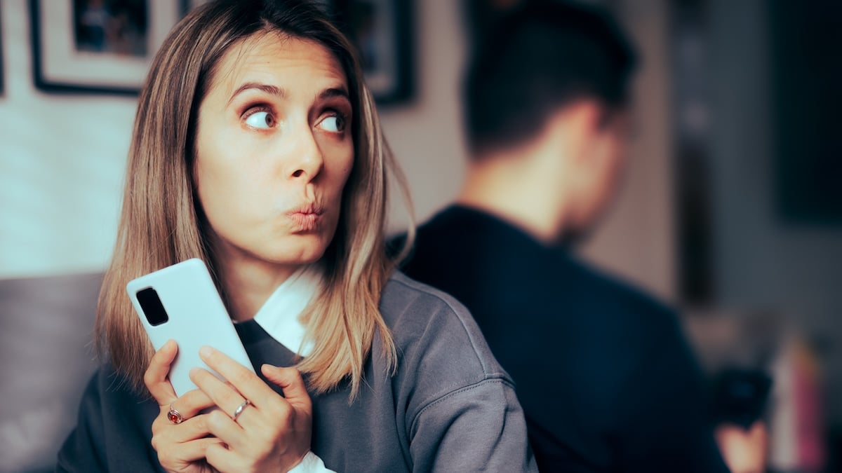 Affäre bei WhatsApp verstecken: Frau steht mit dem Rücken zu einem Mann und hält schuldbewusst ihr Handy in der Hand